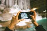 I förgrunden: två händer som håller en mobil och fotar Fontana di Trevi, som visas i bakgrunden.